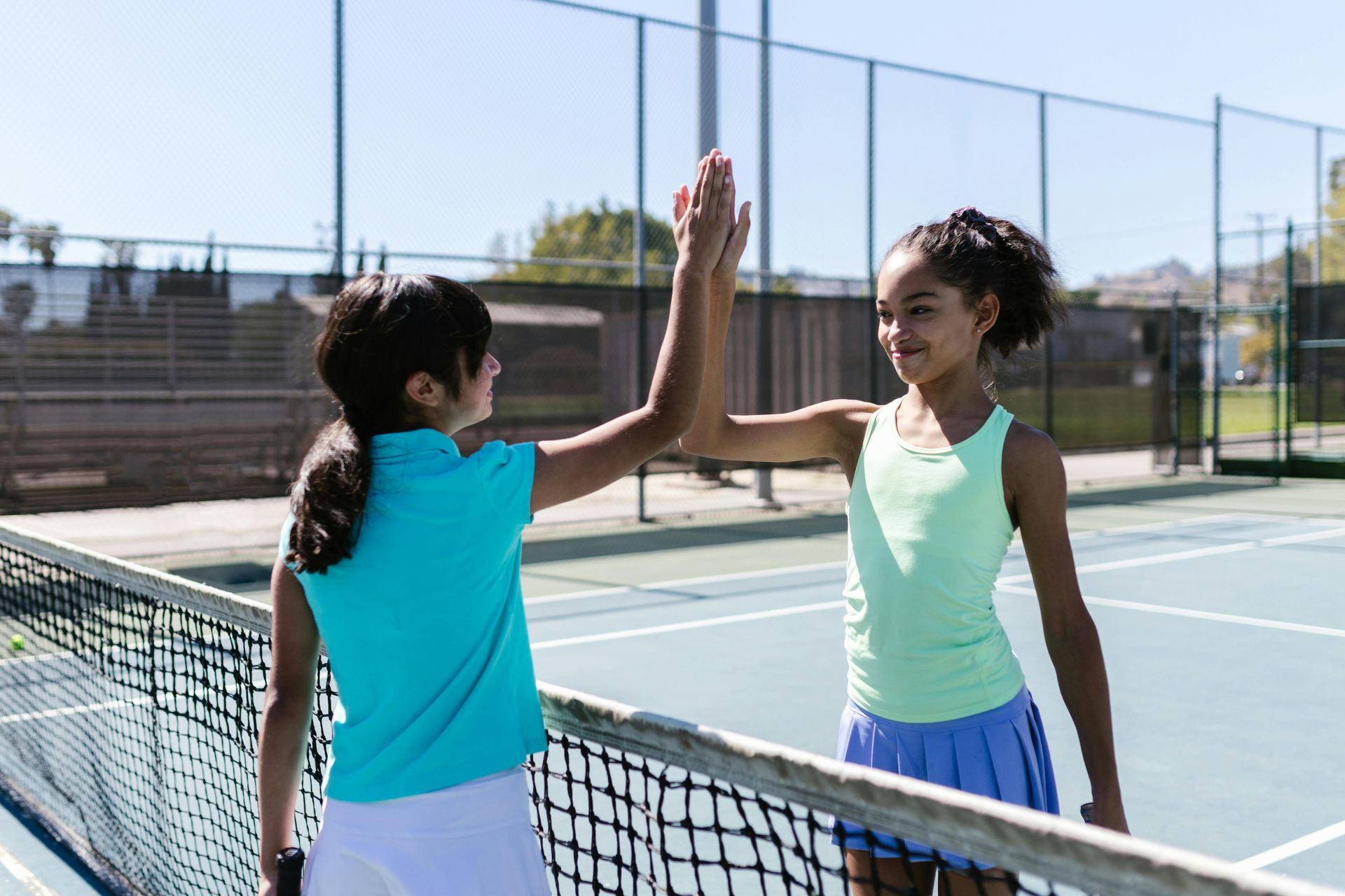 Two girls high-five on a tennis court, standing by the net in bright sportswear.