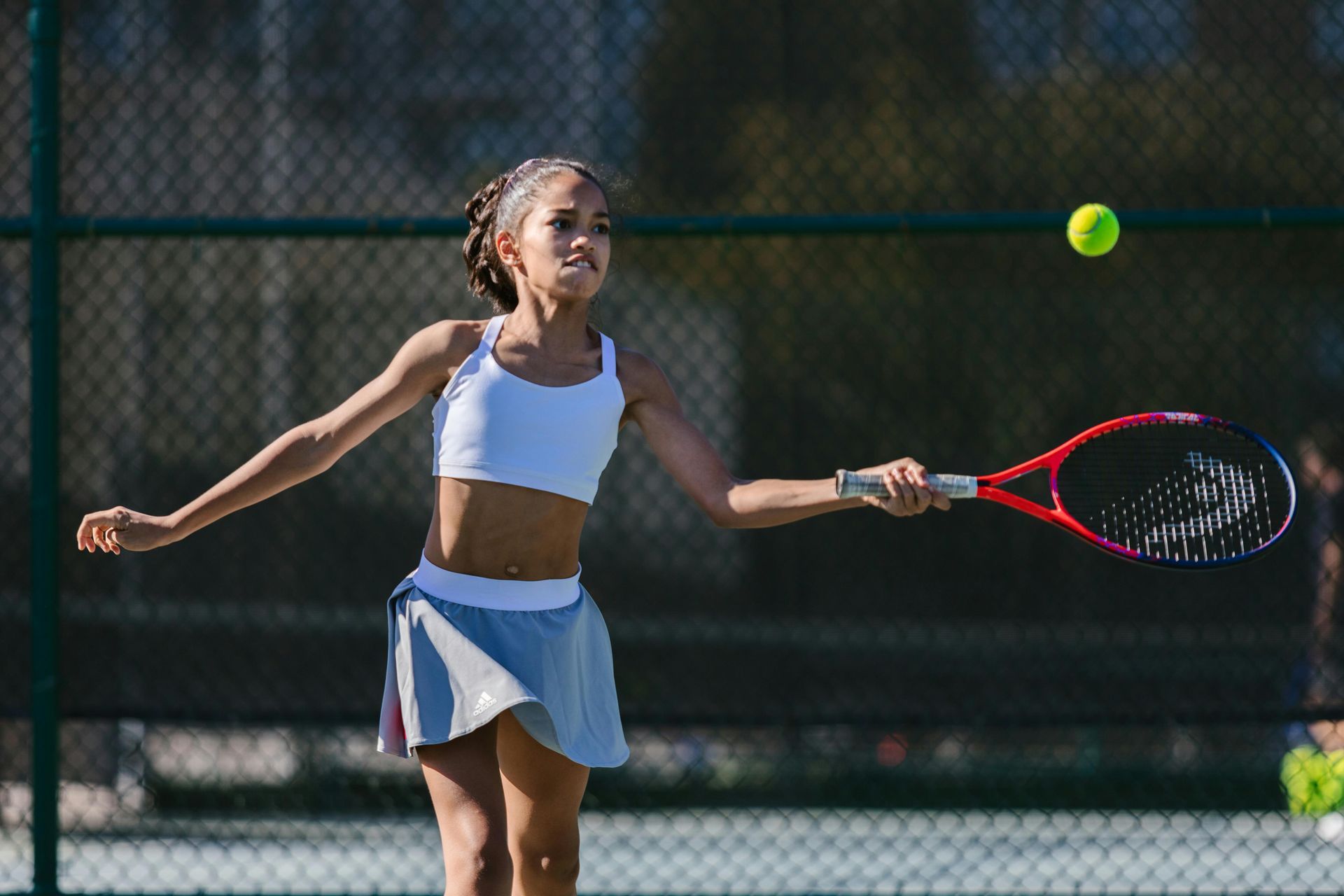 A person in athletic wear swings a red tennis racket at a yellow ball on a fenced tennis court.