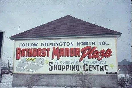 Large roadside sign for Bathurst Manor Plaza shopping centre, with red and black lettering on a snowy day.