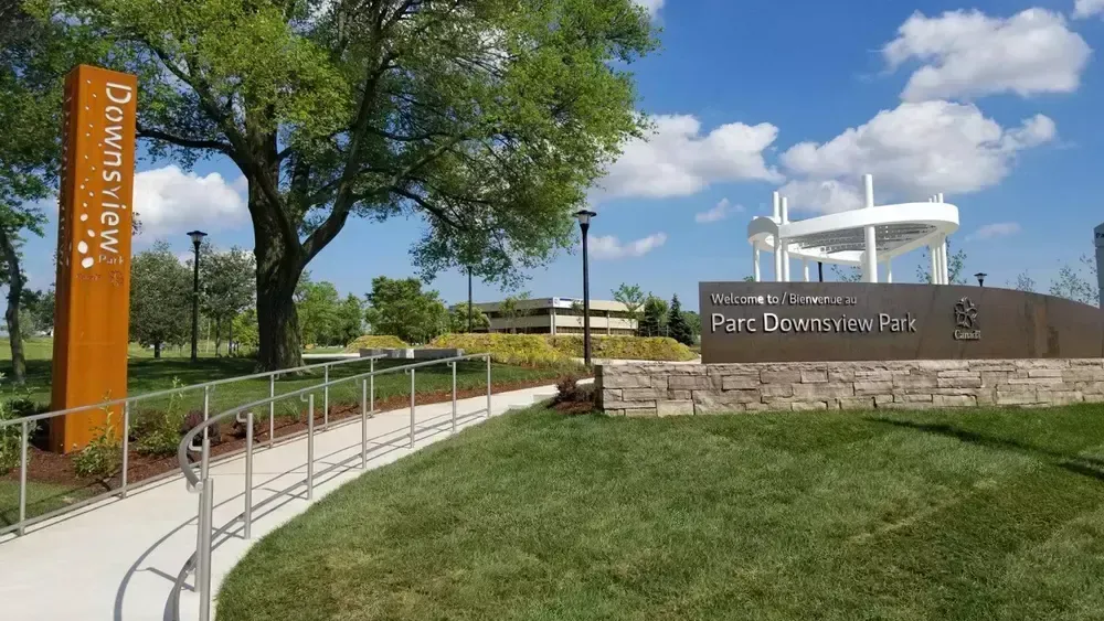 Park entrance with curved sidewalk, stone sign, and white pavilion under blue sky.