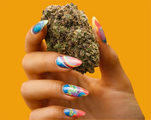Hand holding a large cannabis bud with long, colorful, patterned nails on an orange background