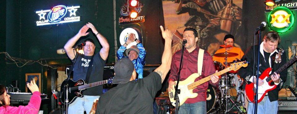 A band performs on stage in a bar, with a crowd member raising their hand. The band members play guitars and drums.