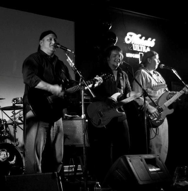 A black and white photo shows a band performing on stage. Three musicians are playing guitars and singing with a 