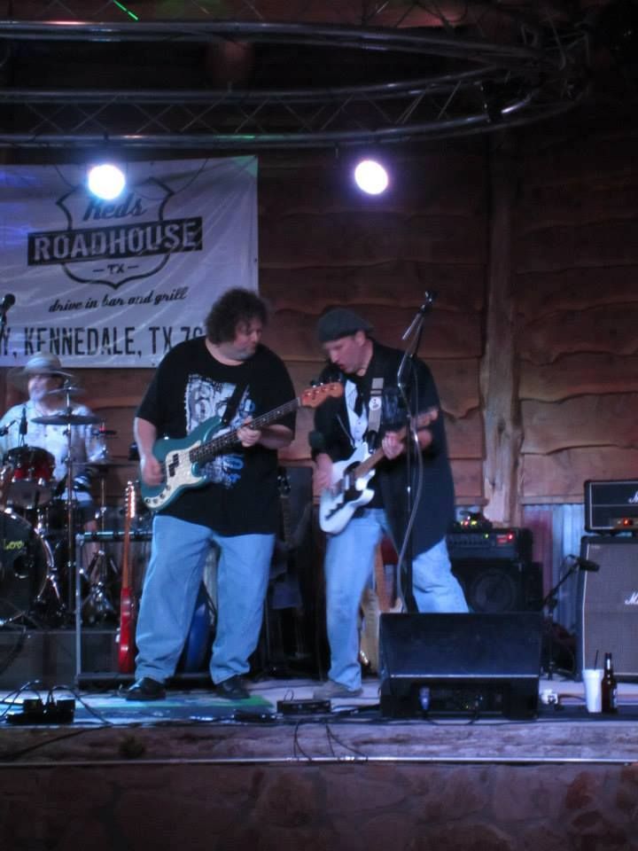 Band performing on stage at a roadhouse. Two guitarists play while a drummer sits behind them. Banner reads 