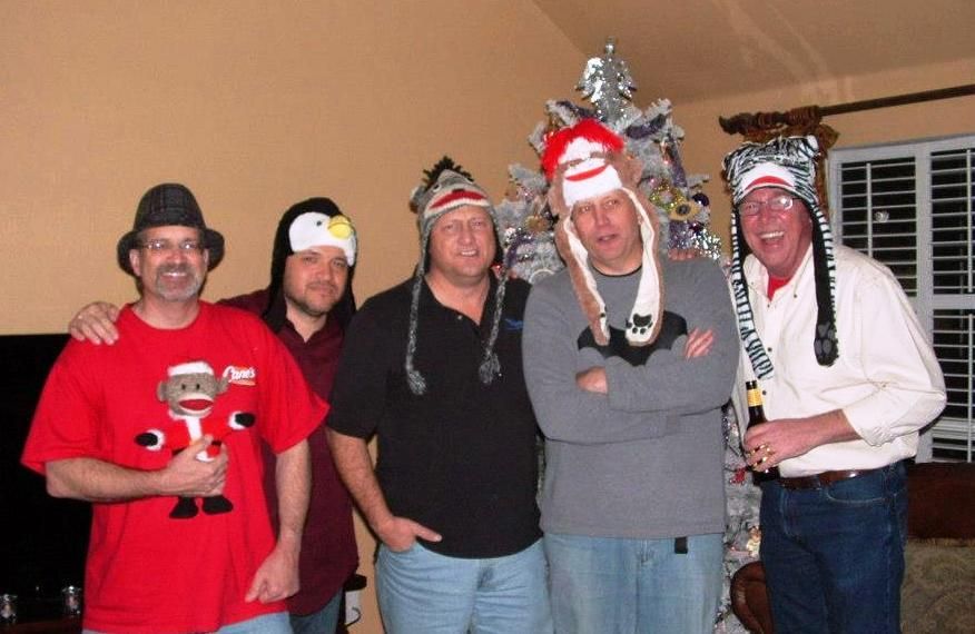 Five men wearing novelty hats, posing for a photo in front of a Christmas tree. One man holds a beer, and another has his arms crossed.