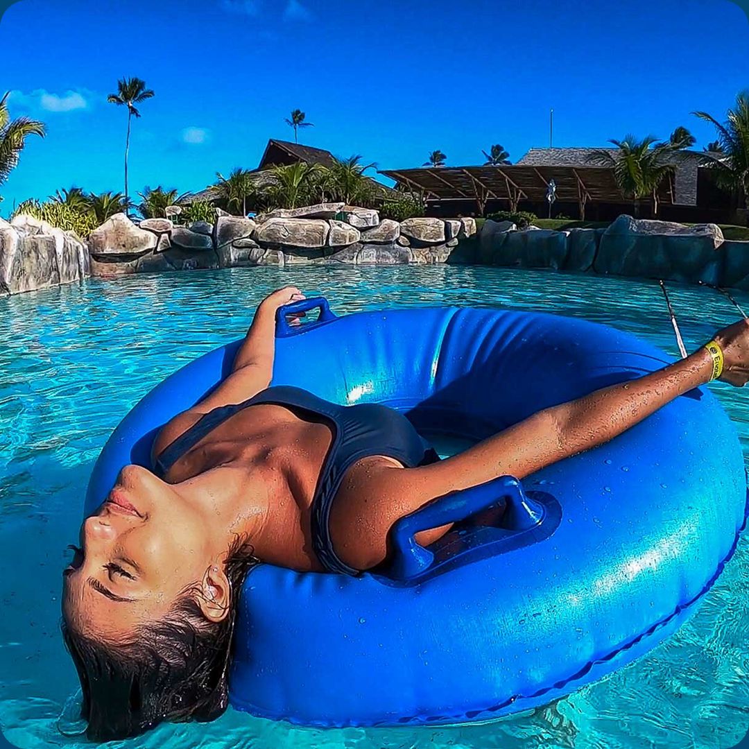 Close-up de uma mulher relaxando de biquíni escuro deitada em uma boia azul circular em um rio lento de água azul. Ao fundo, coqueiros, rochas paisagísticas e a arquitetura do resort all inclusive Enotel Porto de Galinhas sob um céu azul ensolarado. Uma representação de relaxamento nas férias de dezembro.