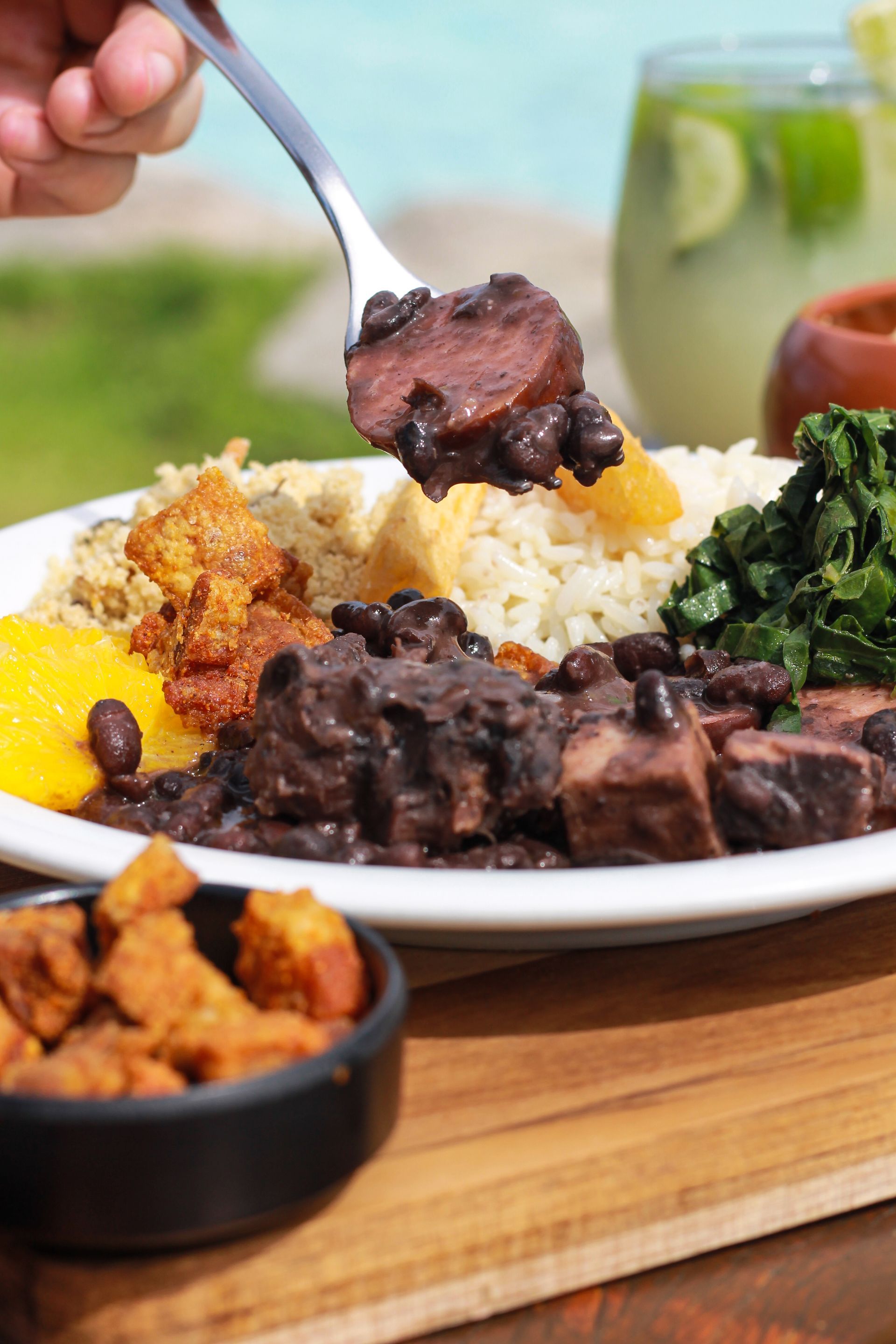 Close-up de um prato de feijoada brasileira com arroz branco, couve refogada, farofa e torresmo. Um garfo levanta uma rodela de linguiça suculenta coberta com feijão preto.