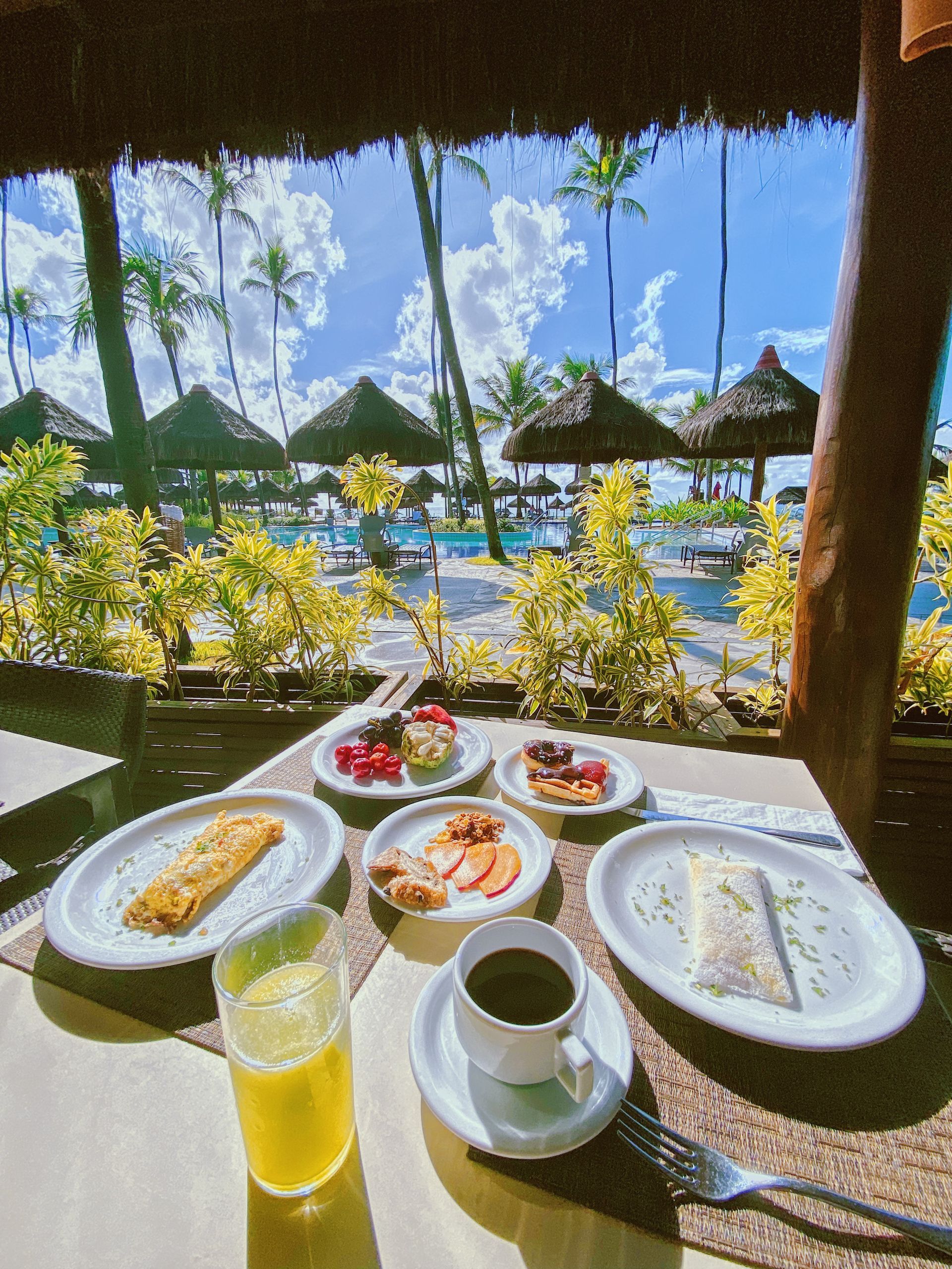Mesa de café da manhã com omelete, tapioca e frutas. Ao fundo, vista tropical com plantas, coqueiros e quiosques na área da piscina do resort.