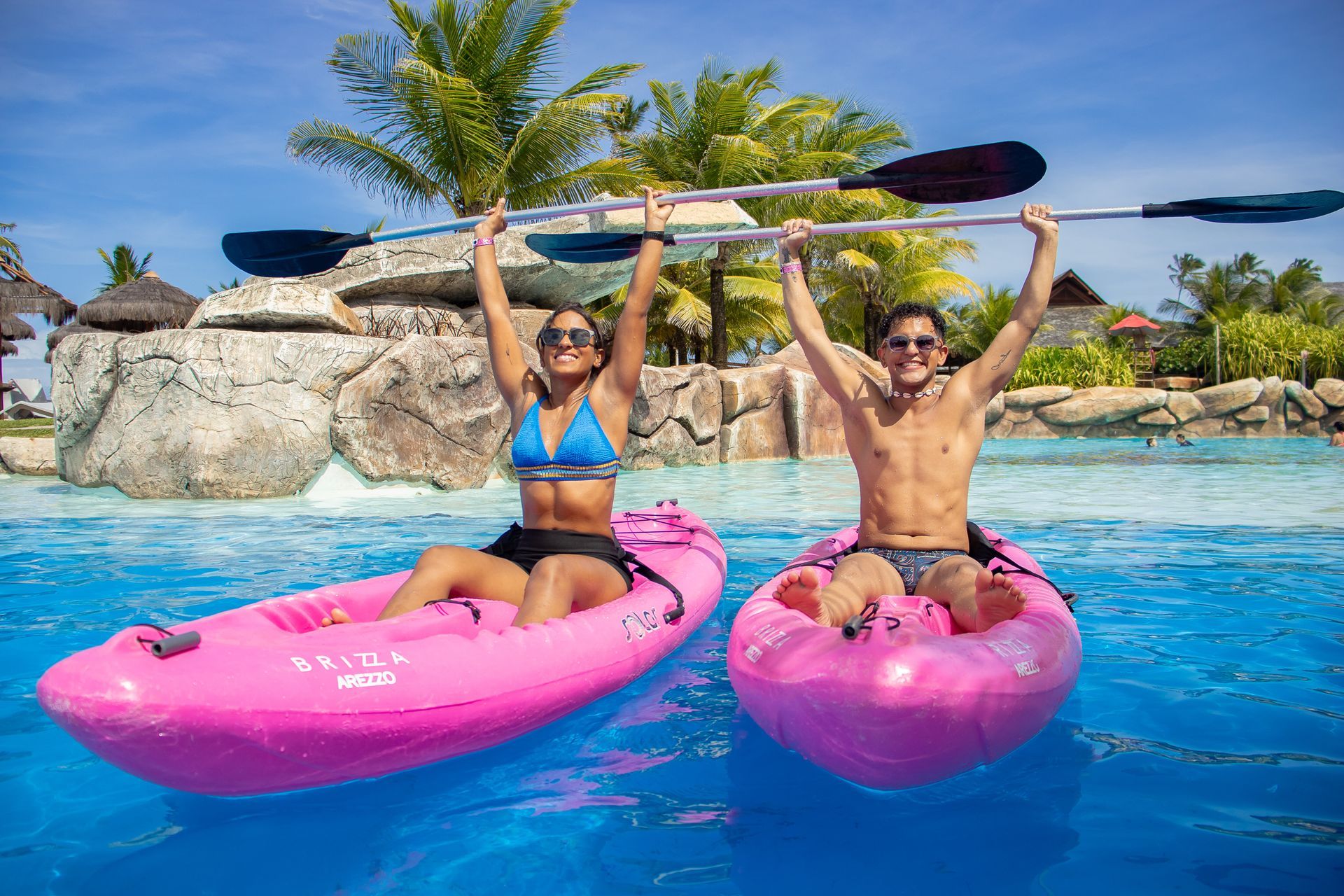 Casal sorridente em caiaques rosa na piscina do resort Enotel. Eles levantam os remos em clima de diversão sob o sol de Porto de Galinhas.