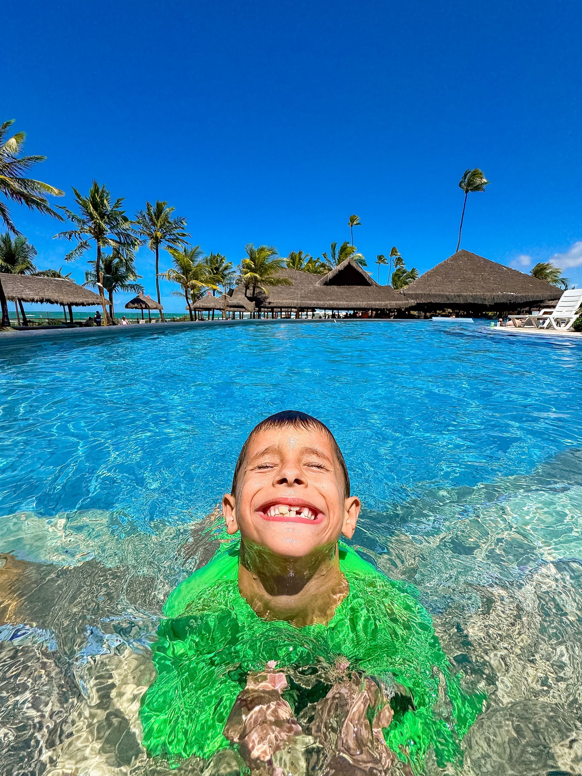 Menino sorridente mergulhando na piscina azul do resort. Fundo com coqueiros e quiosques sob céu aberto.