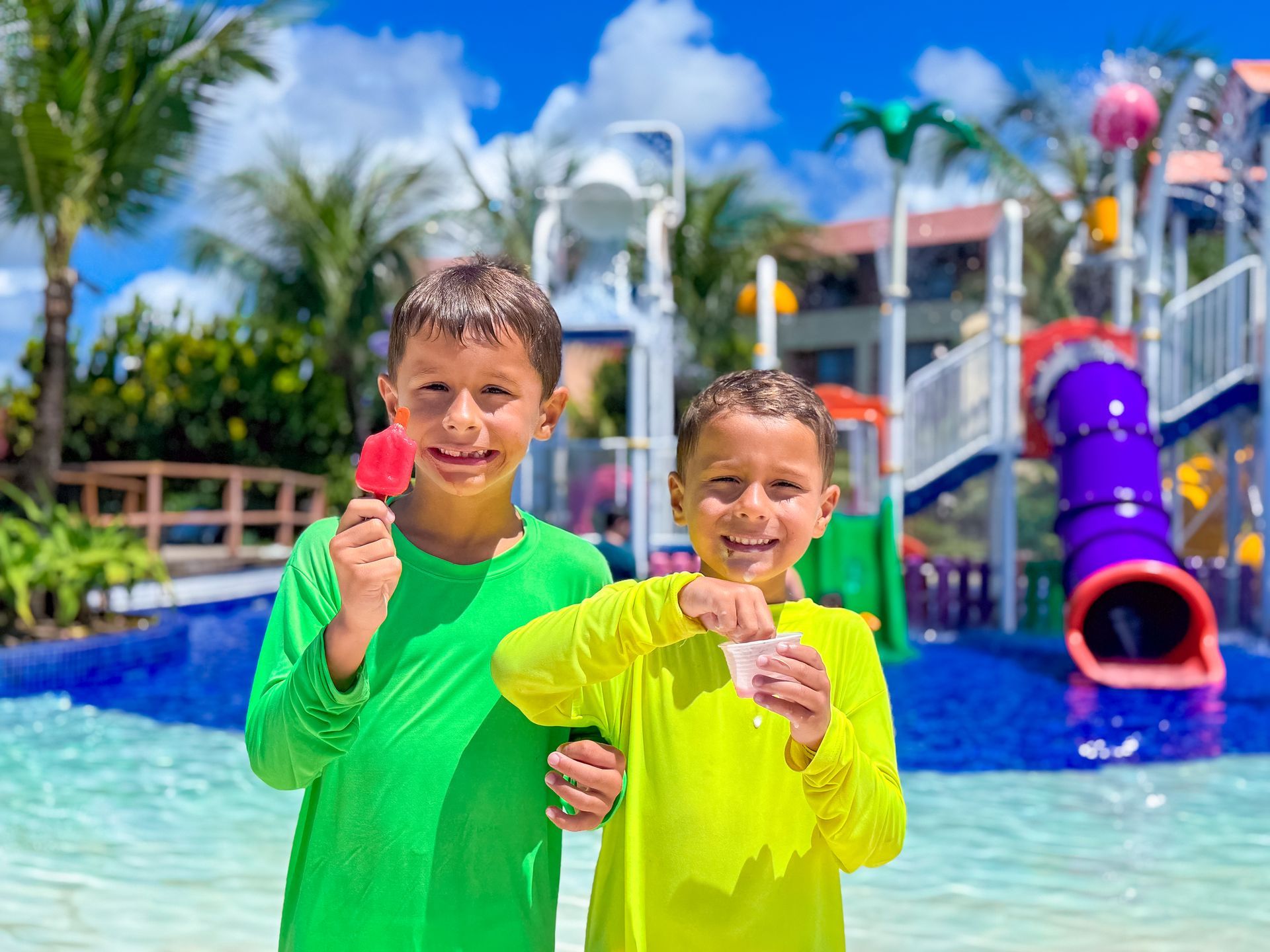 Dois meninos sorridentes segurando picolé e sorvete em frente ao parque aquático do Enotel. Diversão refrescante no sistema all inclusive.
