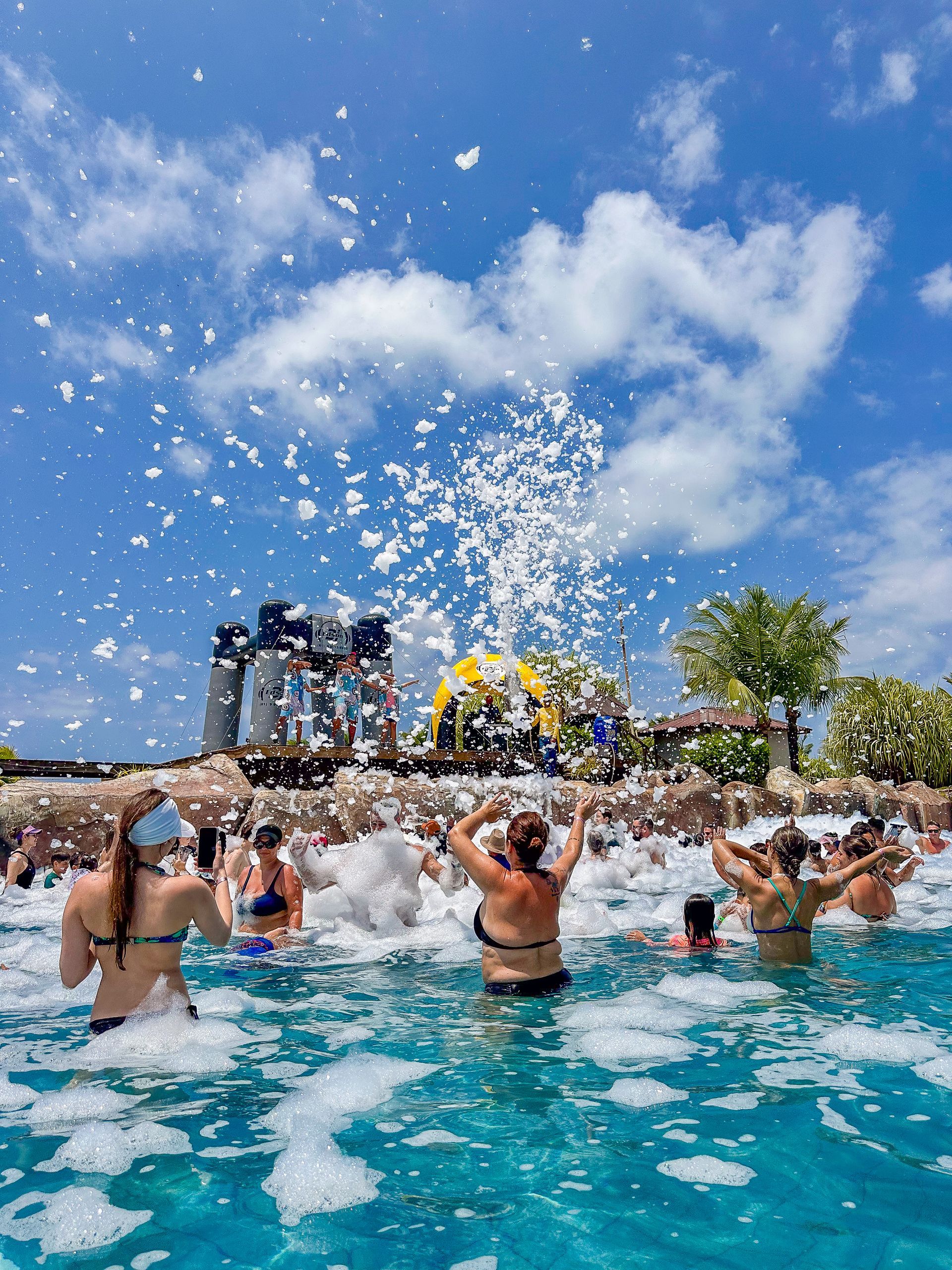 Pessoas celebrando em uma festa na espuma dentro de uma piscina de águas azuis. Há jatos de espuma branca lançados ao ar sob um céu azul ensolarado com coqueiros ao fundo.