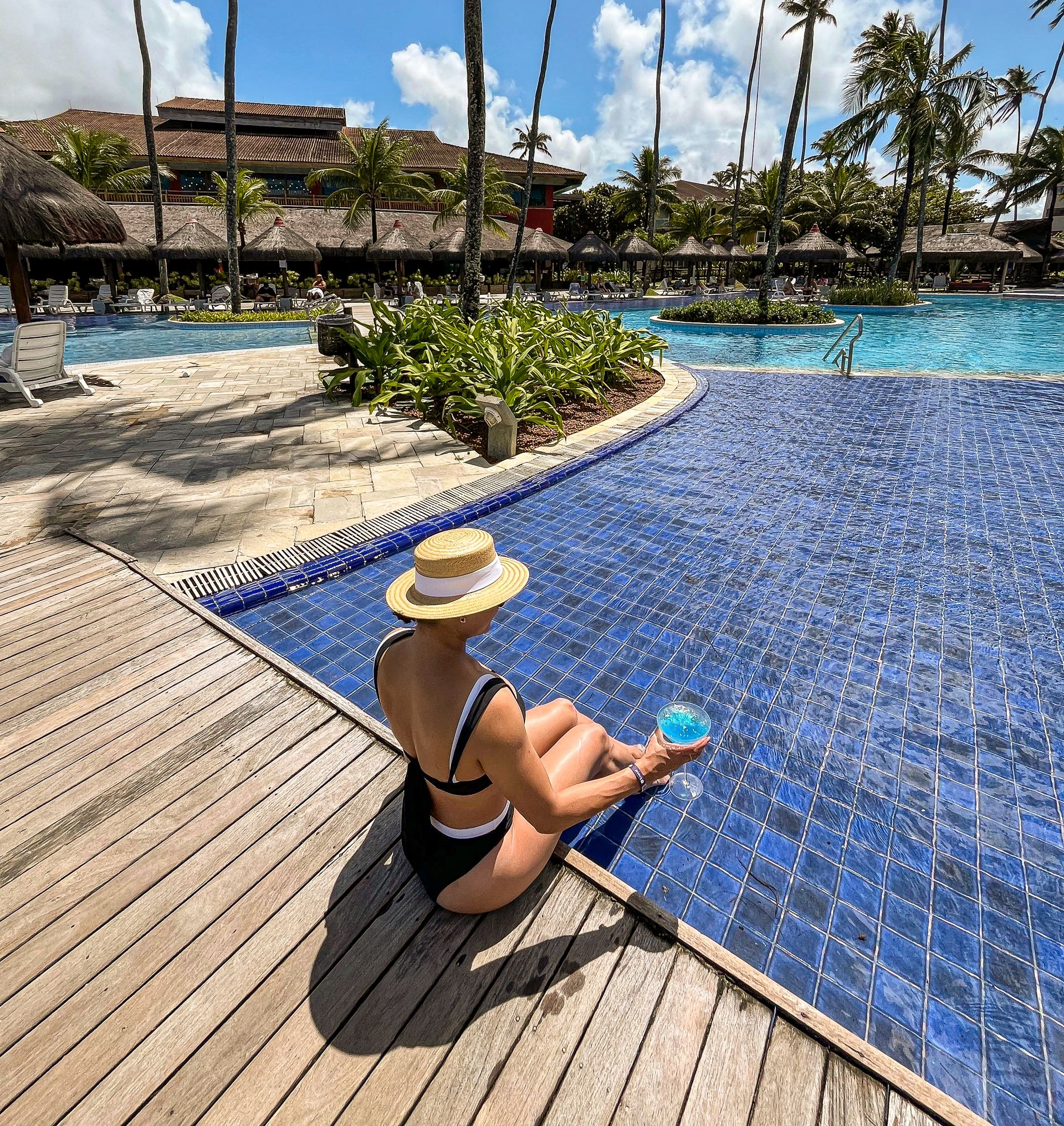 Mulher de chapéu de palha sentada de costas na borda de uma piscina de azulejos azuis, segurando um drink azul vibrante. Ao fundo, coqueiros altos e a estrutura do hotel.