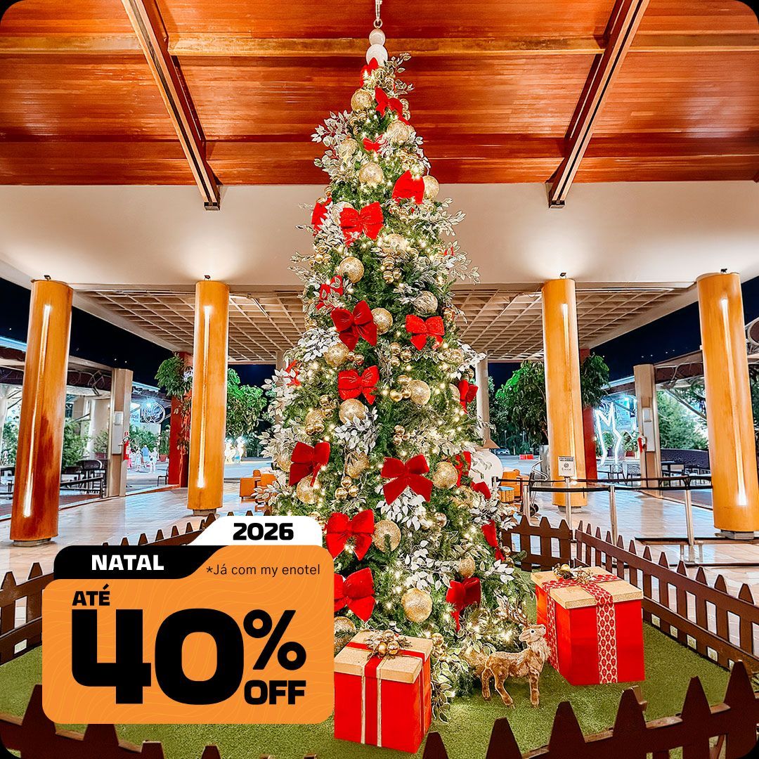 Grande árvore de Natal iluminada, decorada com laços vermelhos e bolas douradas, junto a caixas de presentes no resort Enotel. A imagem exibe o selo da promoção My Black com até 40% de desconto para reservas de Natal em 2026.
