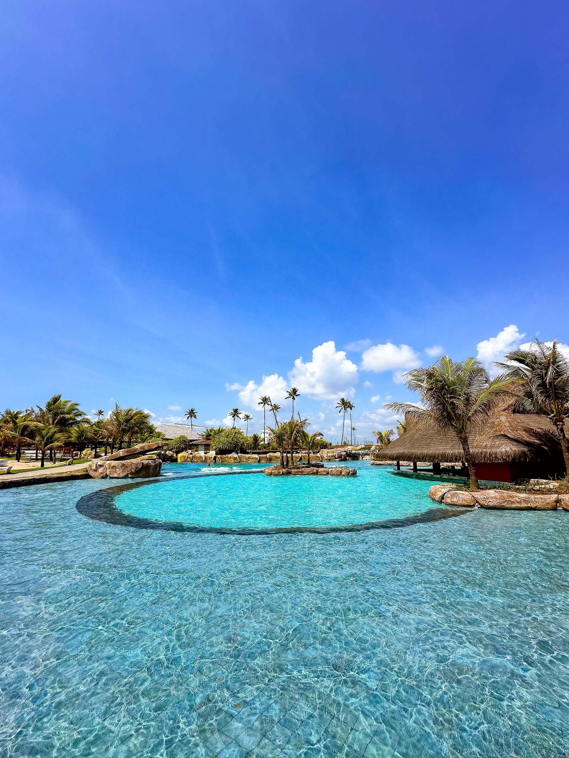 Vista da piscina circular azul do resort Enotel. Cercada por coqueiros e quiosques rústicos sob um céu azul ensolarado com nuvens.