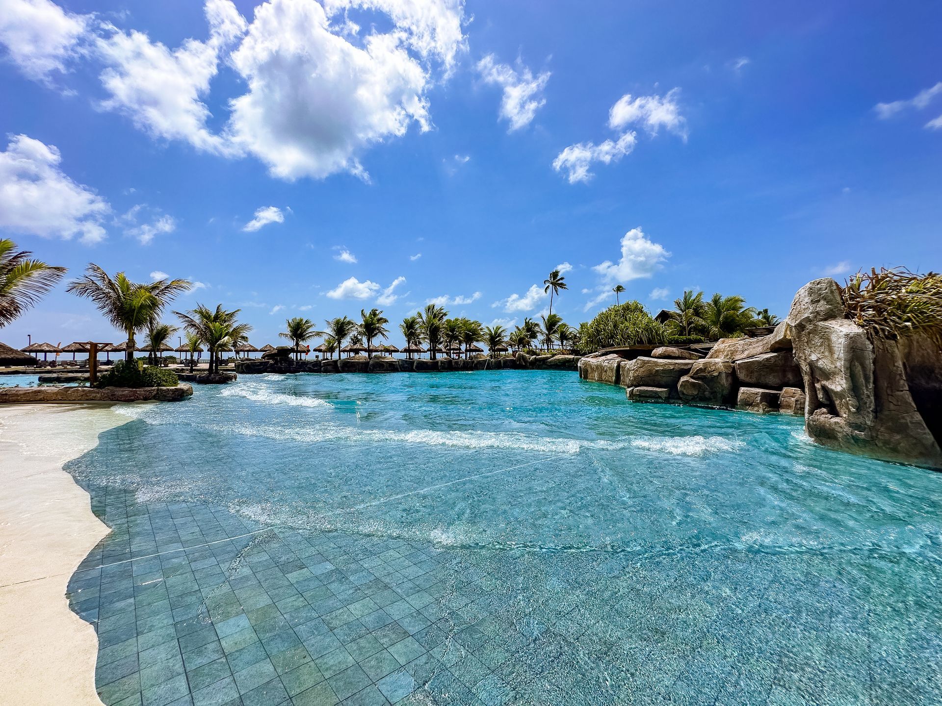 Vista ampla de uma piscina de ondas cristalina cercada por areia branca, rochas decorativas e muitos coqueiros sob um céu com nuvens esparsas.