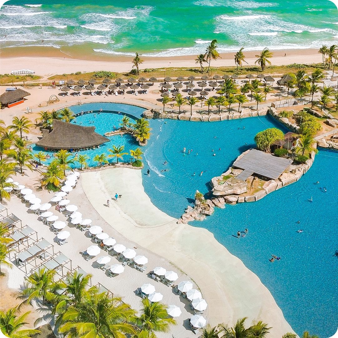 Vista aérea da ampla piscina com praia artificial, águas cristalinas e dezenas de guarda-sóis brancos no resort Enotel Porto de Galinhas, com o mar ao fundo. O ambiente ideal para relaxar e aproveitar os primeiros dias do ano durante a temporada de Réveillon.