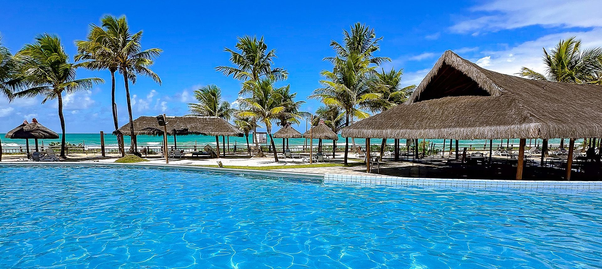 Área de descanso com espreguiçadeiras e quiosques de palha integrados à grande piscina azul do resort Enotel Porto de Galinhas
