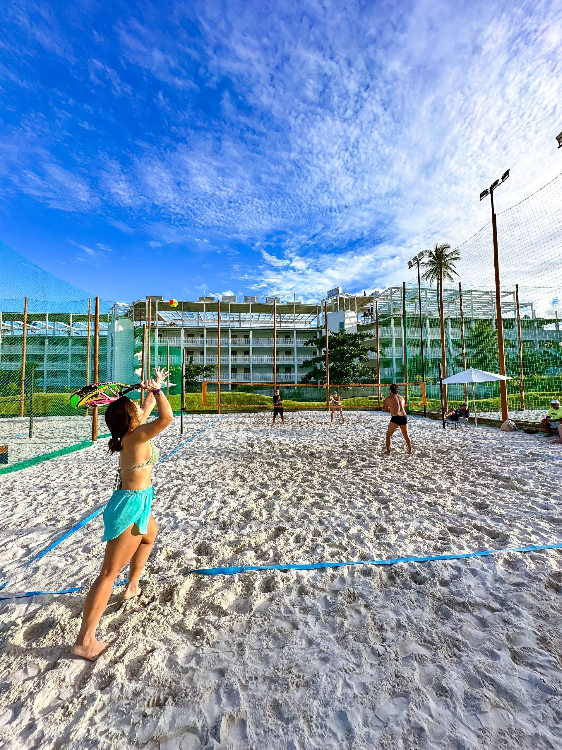 Uma mulher sacando a bola em uma quadra de areia branca de beach tennis. Ao fundo, a rede de proteção, coqueiros e o prédio do resort sob um céu de fim de tarde.