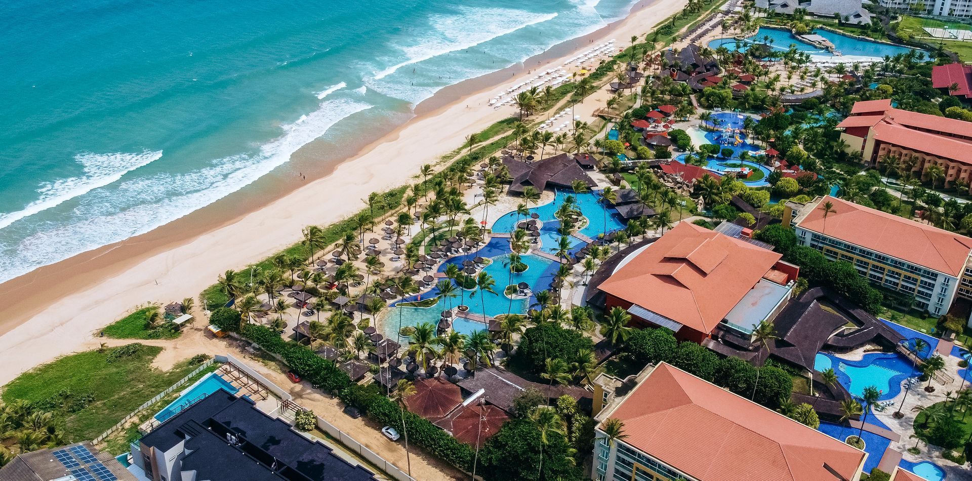 Vista aérea de um resort de luxo à beira-mar com piscinas grandes, coqueiros e praia de areia branca.