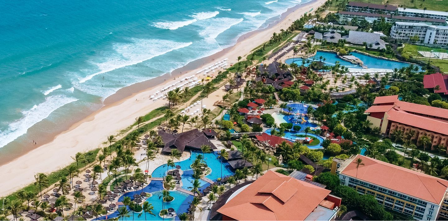 Vista aérea de um resort de luxo à beira-mar com piscinas grandes, coqueiros e praia de areia branca.