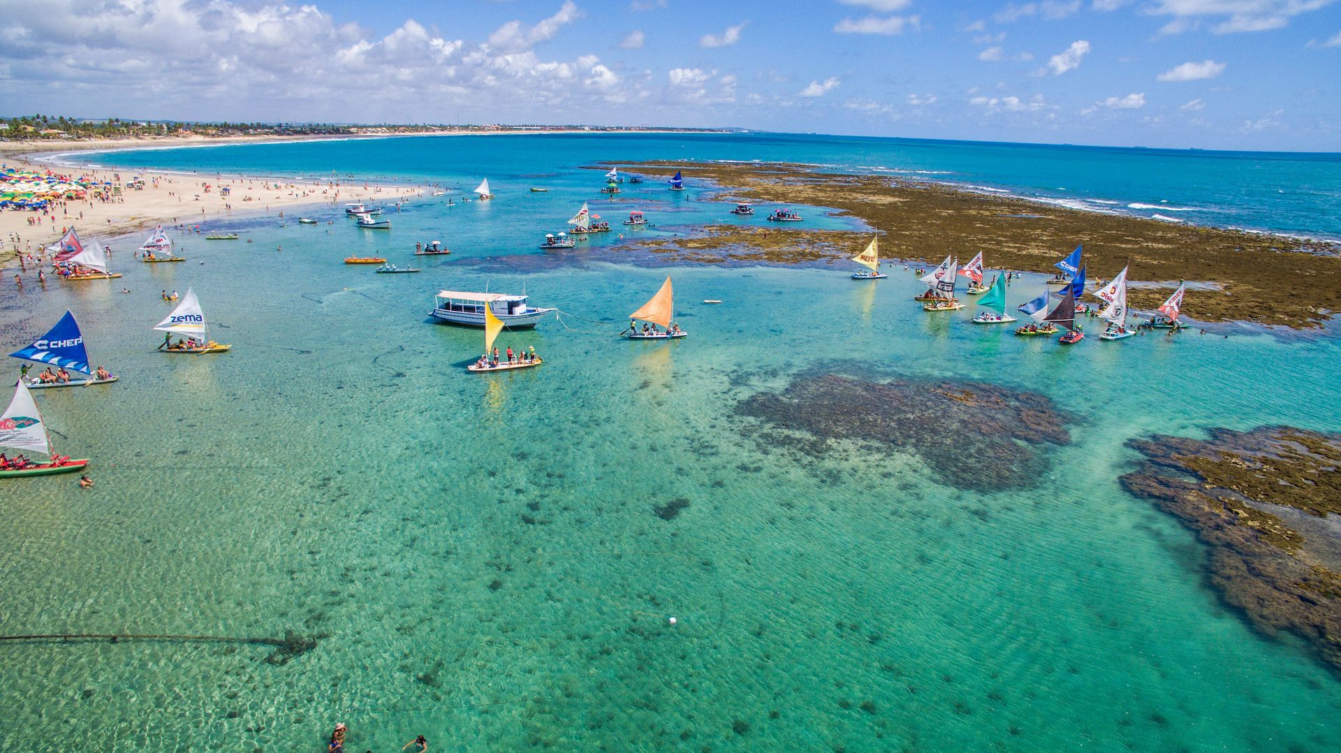 Fotografia aérea das piscinas naturais de águas turquesas e recifes de corais. Diversas jangadas coloridas com velas triangulares navegam próximas à costa de areia clara.