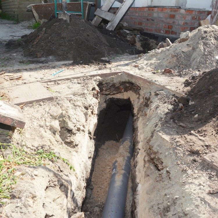 Open trench in a dirt yard exposing a large black pipe near a brick wall and piles of soil
