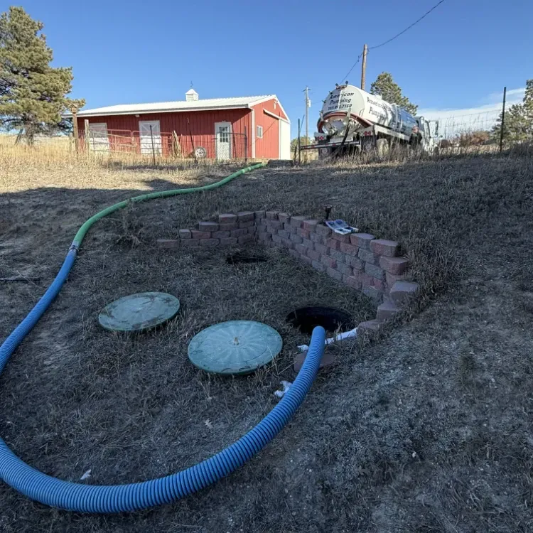 Hoses leading to a basement drain opening in a dry yard near a red house and parked tank truck.