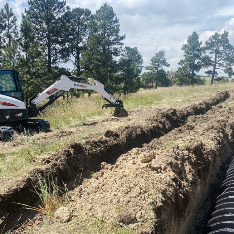 Excavator digging a trench through a grassy field beside a black drainage pipe