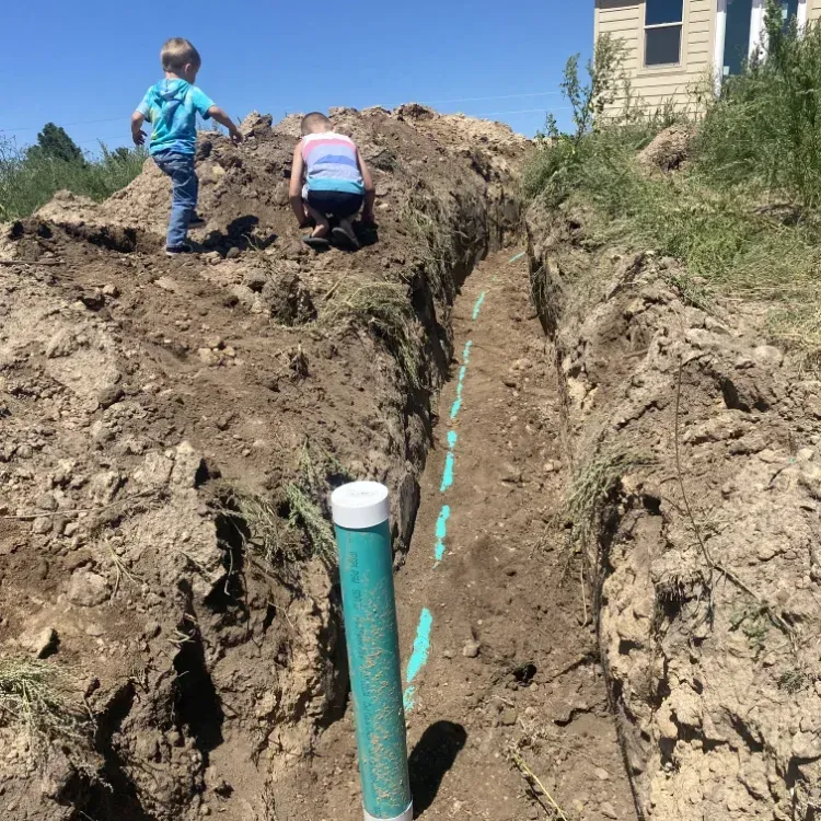 Two children near a trench with a turquoise pipe in a dirt yard beside a house.