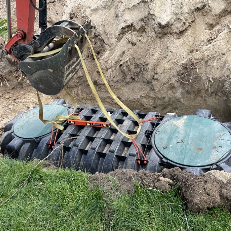 Excavator lifting a black underground tank with yellow straps in a dirt trench beside grass.