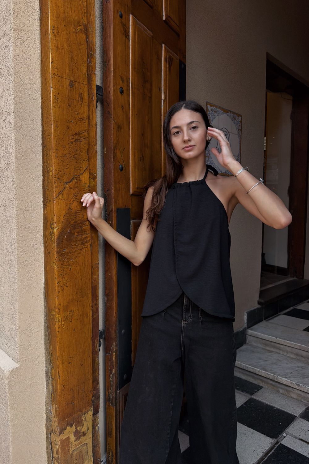 Mujer vestida de negro de pie en el umbral de una puerta, sujetando el marco y tocándose el pelo.
