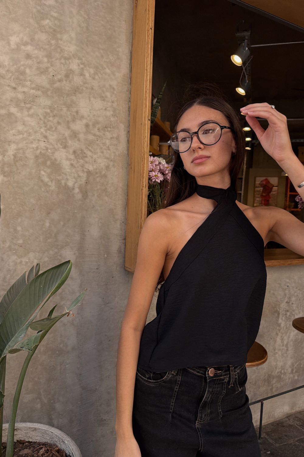 Mujer con top negro sin tirantes y gafas posando junto a la puerta de una cafetería con poca luz.