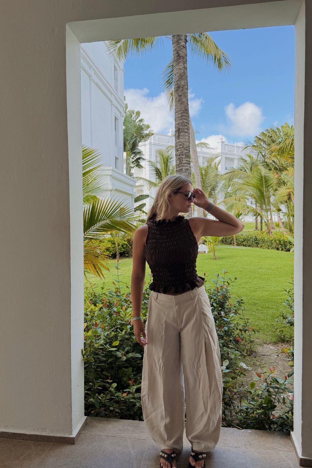 blusa negra y pantalones beige, con una vista a un jardín tropical detrás de ella.