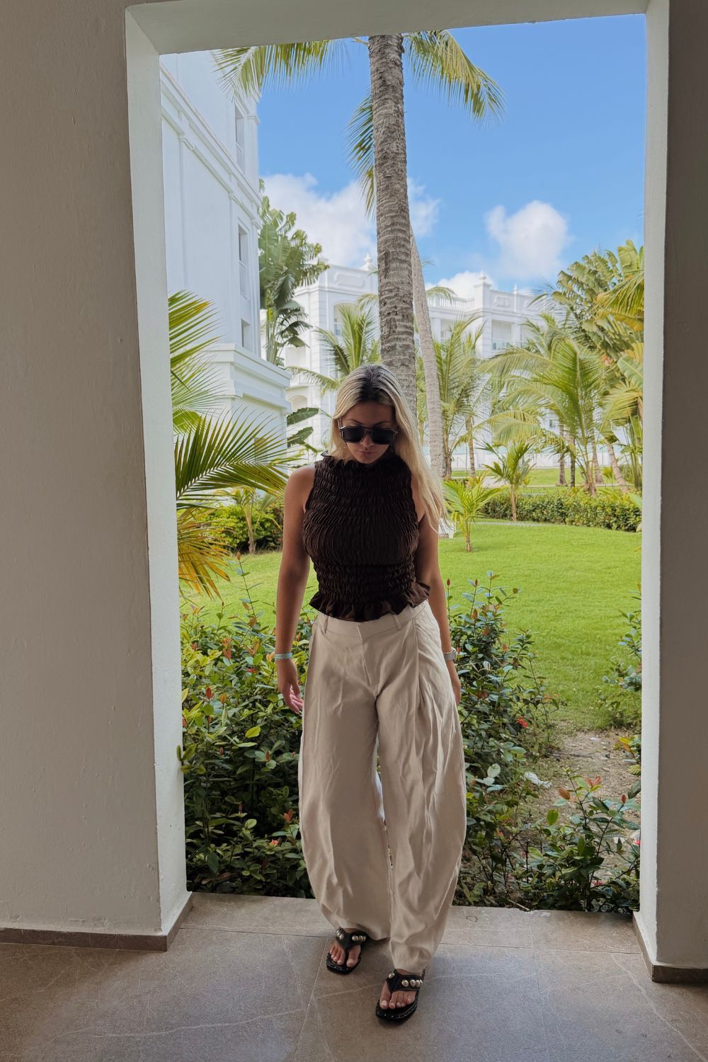 blusa negra y pantalones beige, con una vista a un jardín tropical detrás de ella.