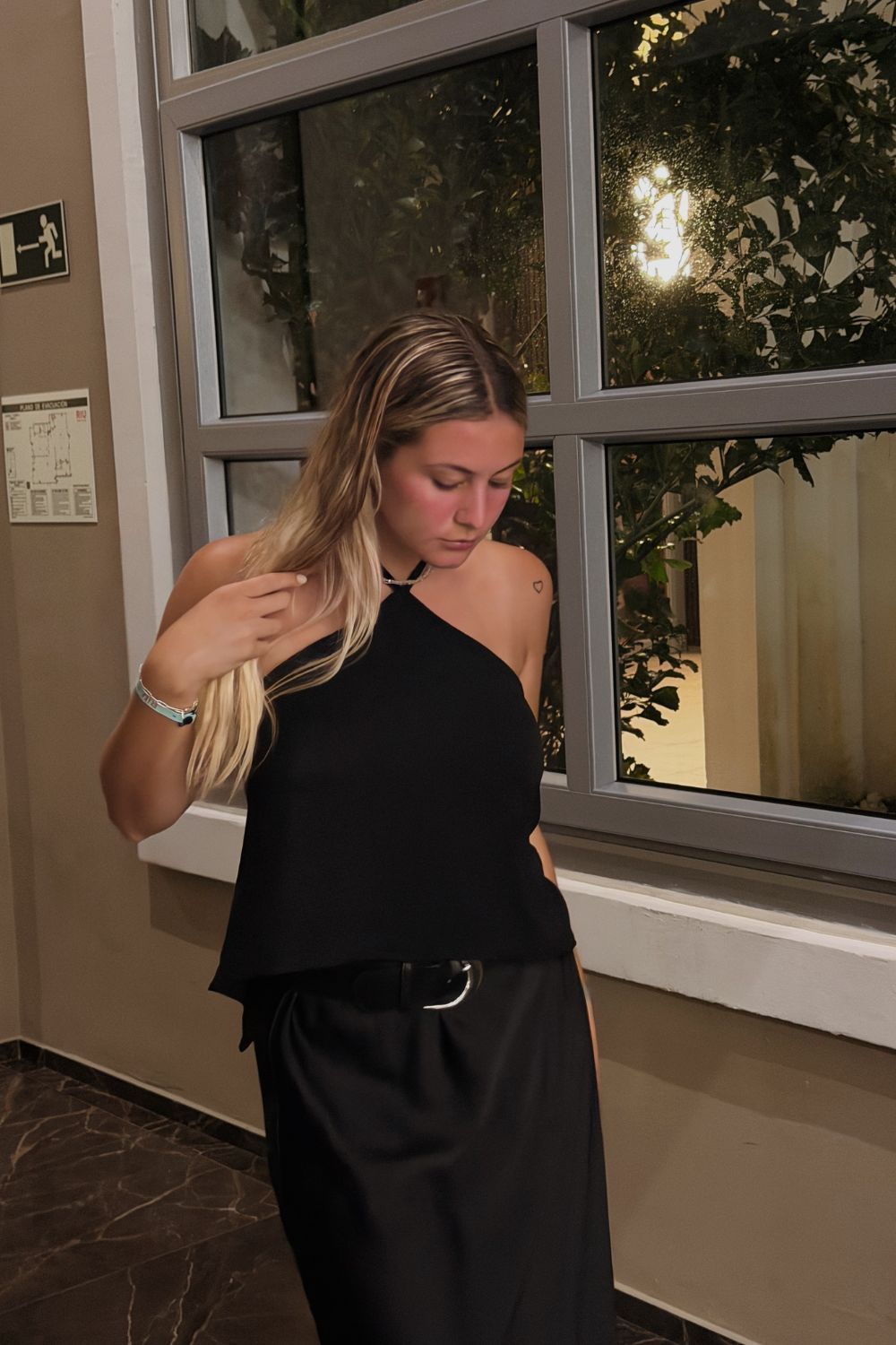 Una persona vestida de negro está de pie junto a una ventana al aire libre, mirando hacia abajo y arreglándose el cabello.