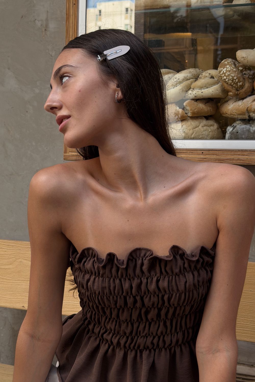 Mujer con un vestido marrón sin tirantes, mirando hacia un lado, sentada afuera cerca de la ventana de una panadería.