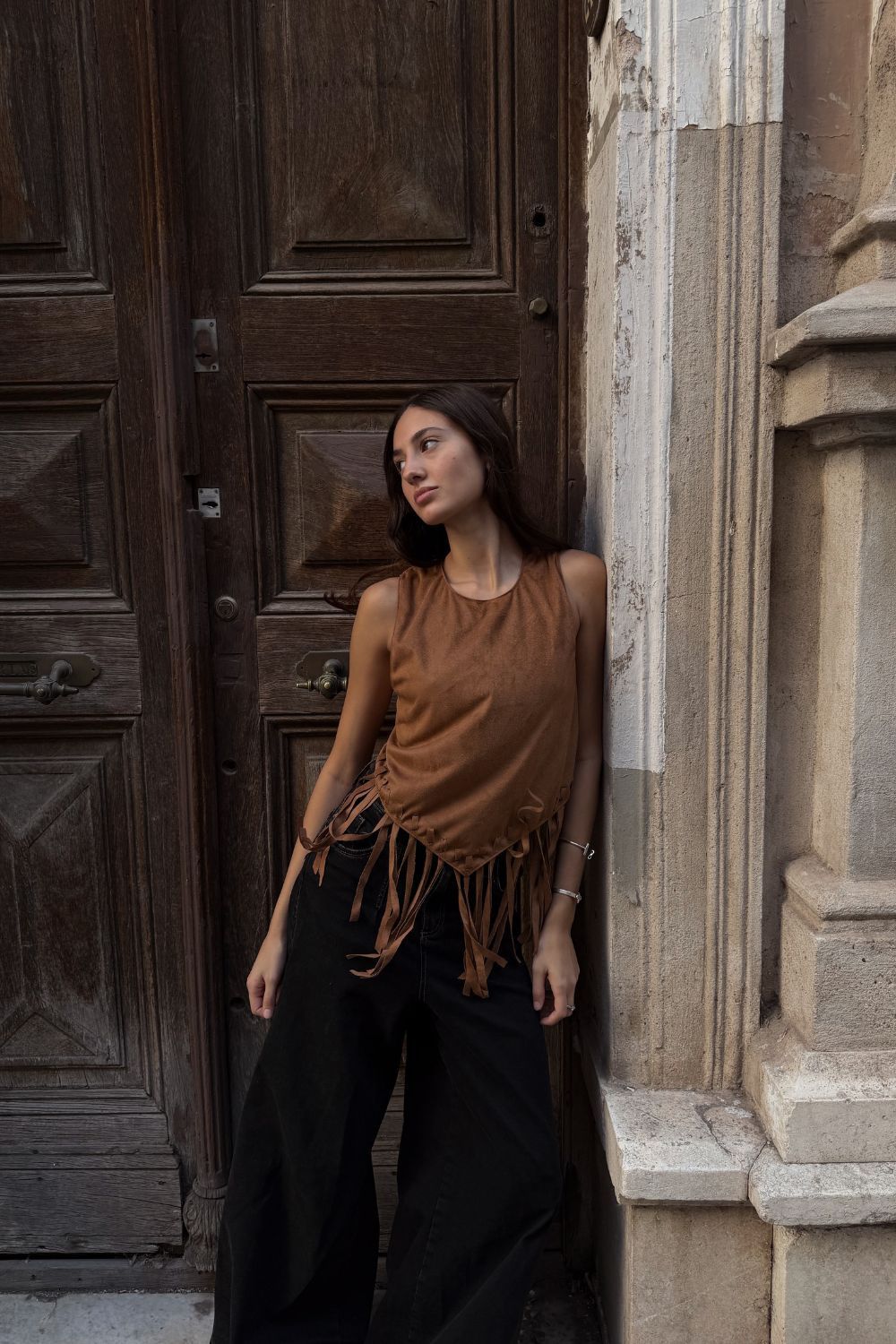 Mujer con una blusa marrón con flecos y pantalones negros apoyada contra un muro de piedra junto a una puerta de madera.