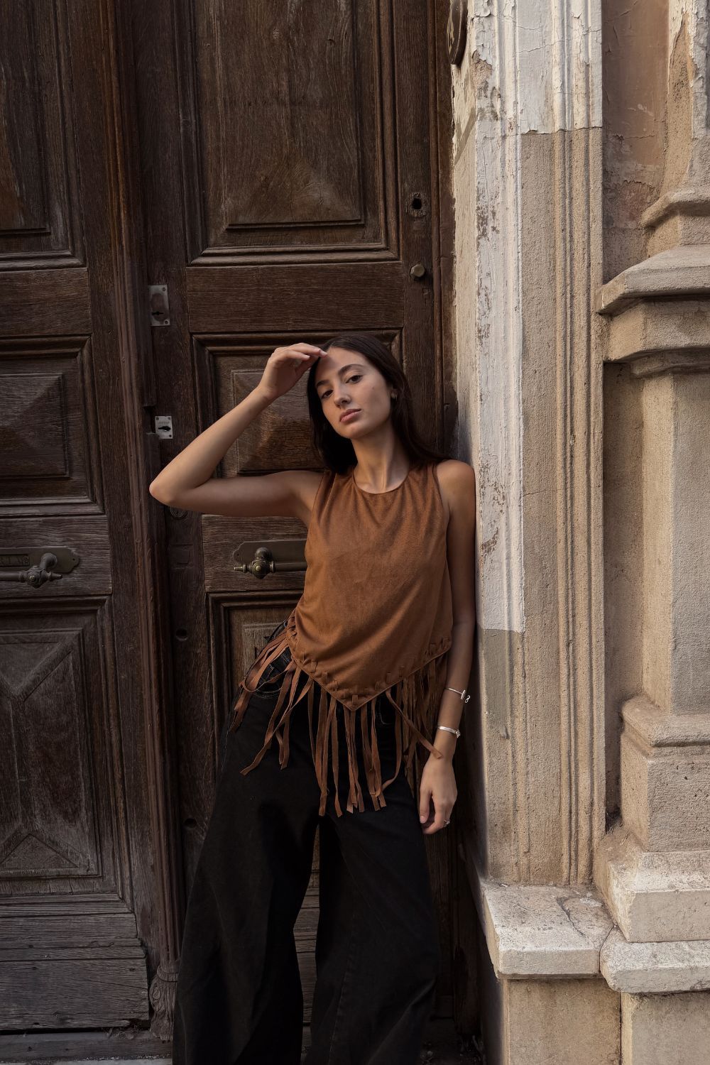 Mujer con una blusa marrón con flecos y pantalones negros apoyada contra un muro de piedra junto a una puerta de madera.