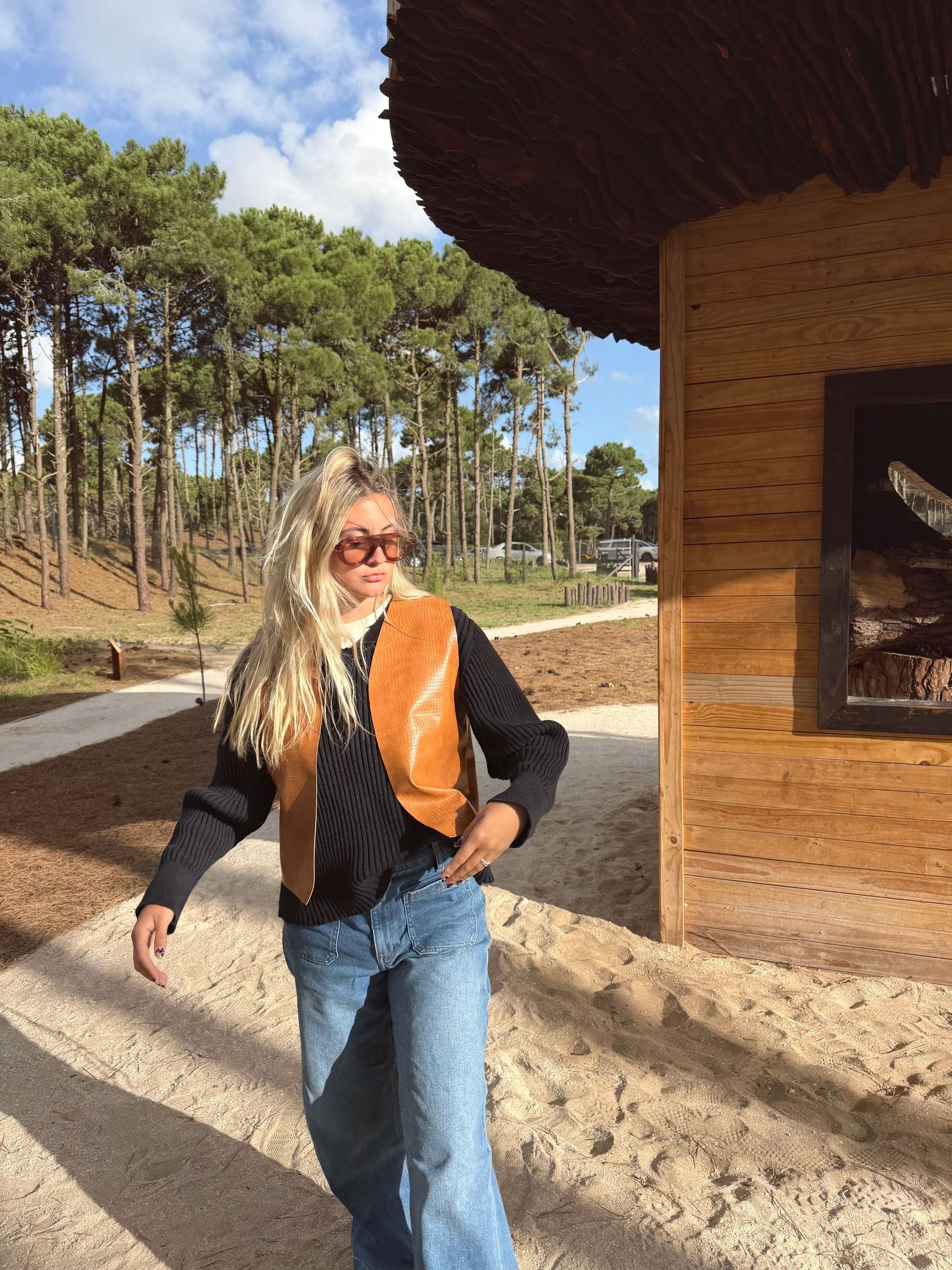 Una persona que viste un chaleco marrón se encuentra junto a una estructura de madera, mirando hacia un sendero forestal.