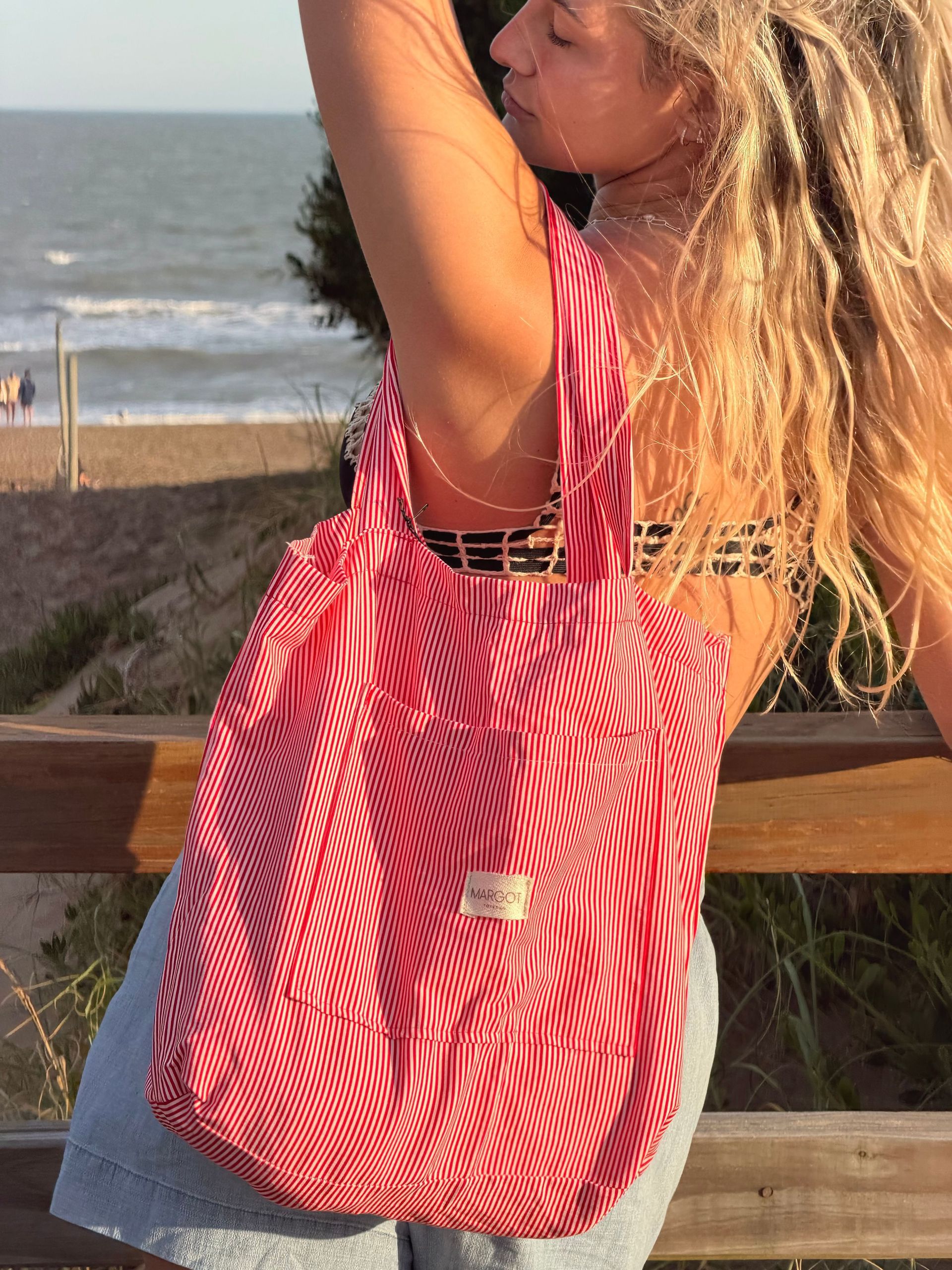 Mujer con cabello rubio, sosteniendo un bolso de rayas rojas y blancas, posando al aire libre cerca del océano.