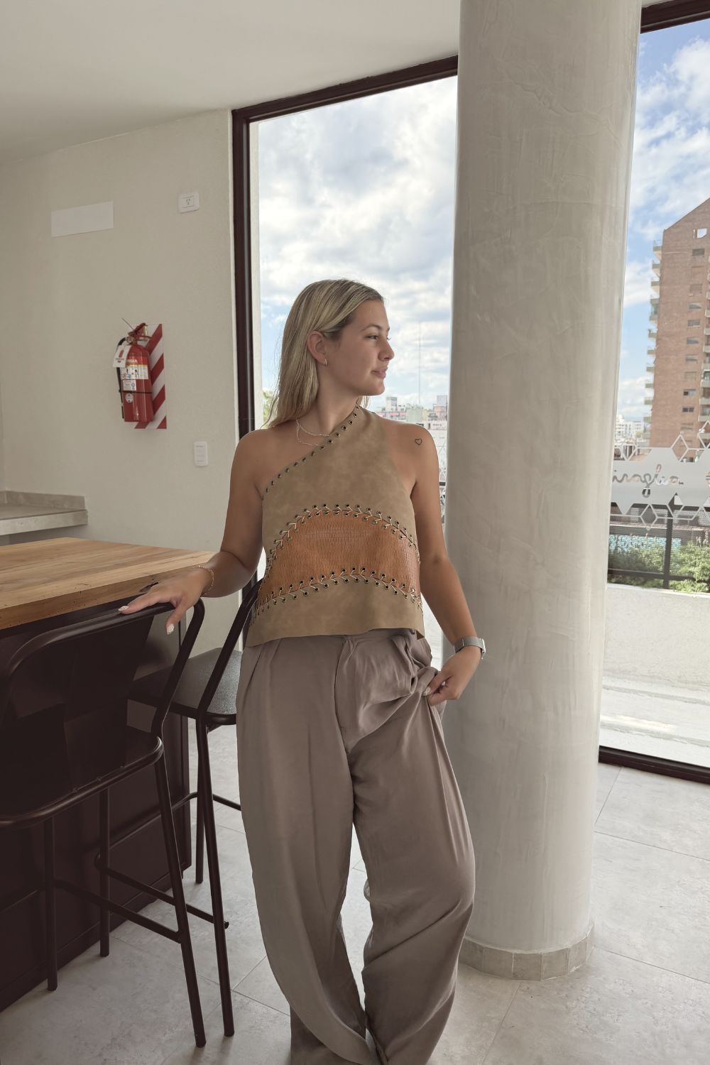 Una mujer vestida con un atuendo beige está de pie junto a una ventana en un luminoso apartamento, mirando hacia afuera.