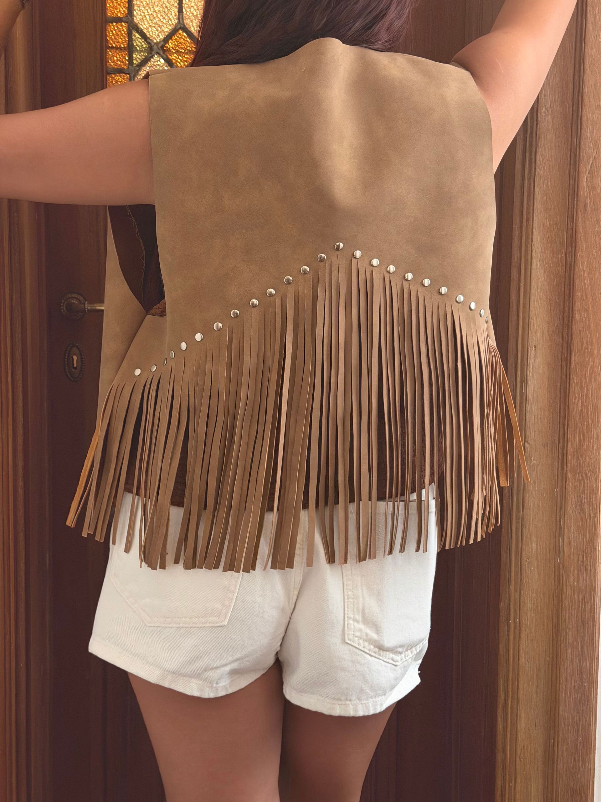 Persona que viste un chaleco de gamuza color canela con flecos sobre pantalones cortos blancos.