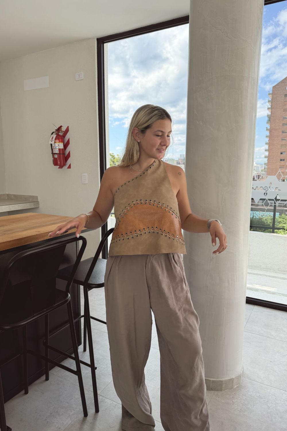 Una mujer vestida con un atuendo beige está de pie junto a una ventana en un luminoso apartamento, mirando hacia afuera.