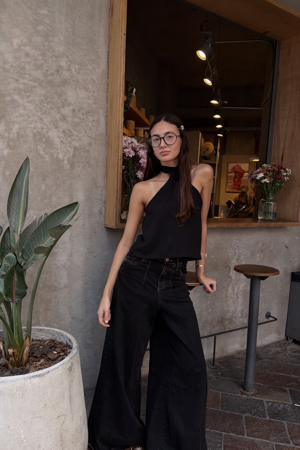 Mujer con top negro sin tirantes y gafas posando junto a la puerta de una cafetería con poca luz.