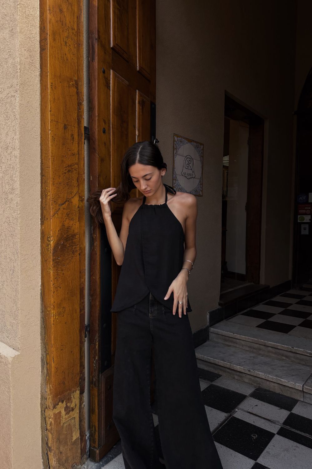 Mujer vestida de negro de pie en el umbral de una puerta, sujetando el marco y tocándose el pelo.