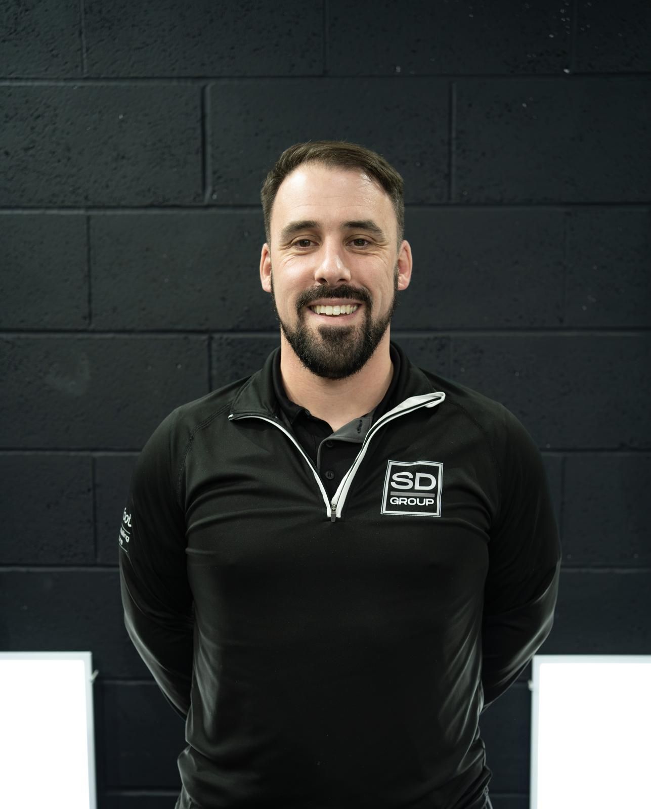 Man with a beard, wearing a black shirt, smiling in front of a black brick wall.