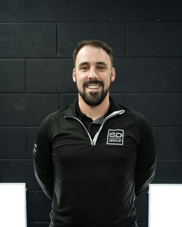 Man with a beard, wearing a black shirt, smiling in front of a black brick wall.