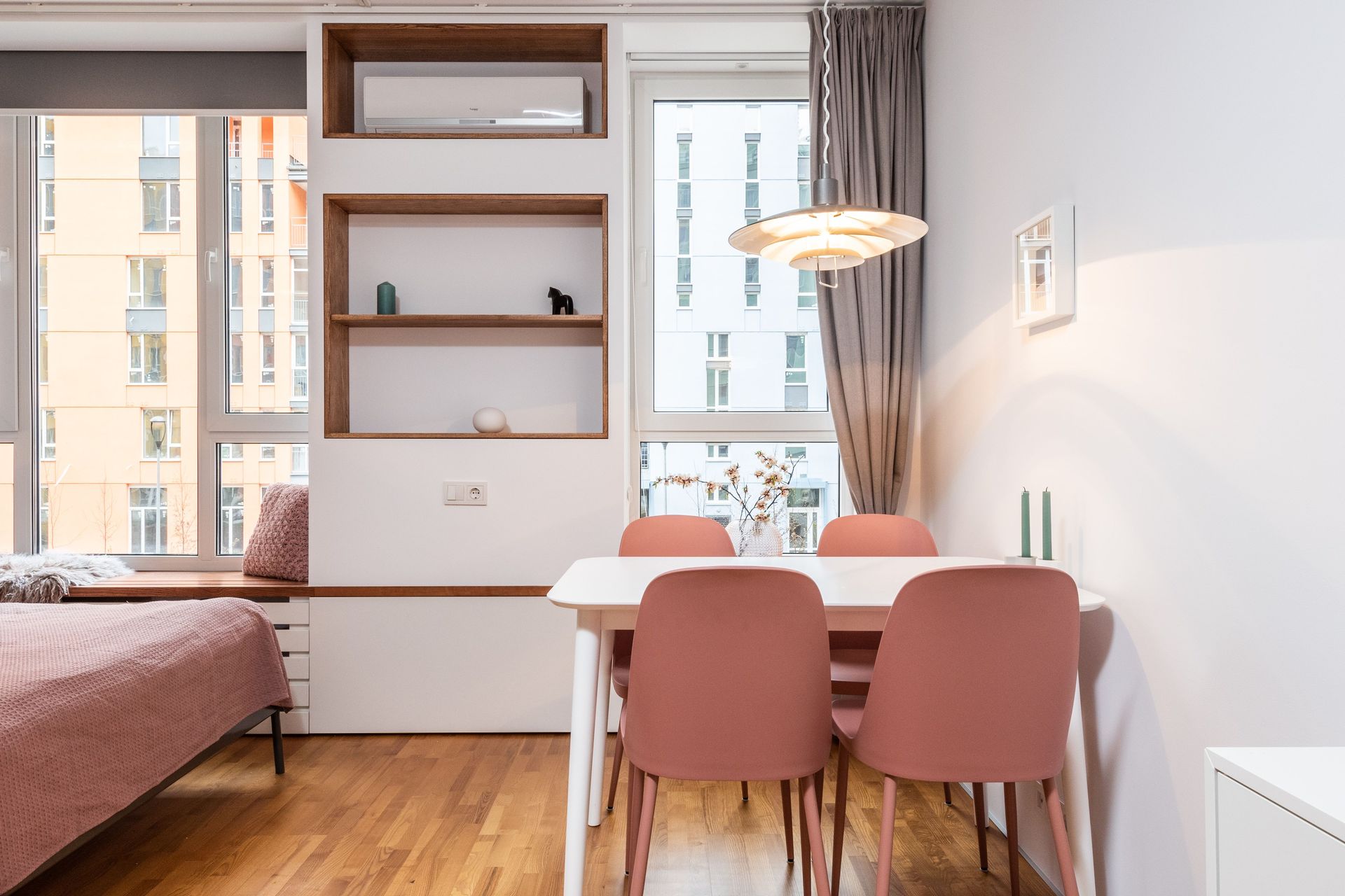 Dormitorio moderno con cama, mesa de comedor y sillas rosas, estanterías empotradas y grandes ventanales.