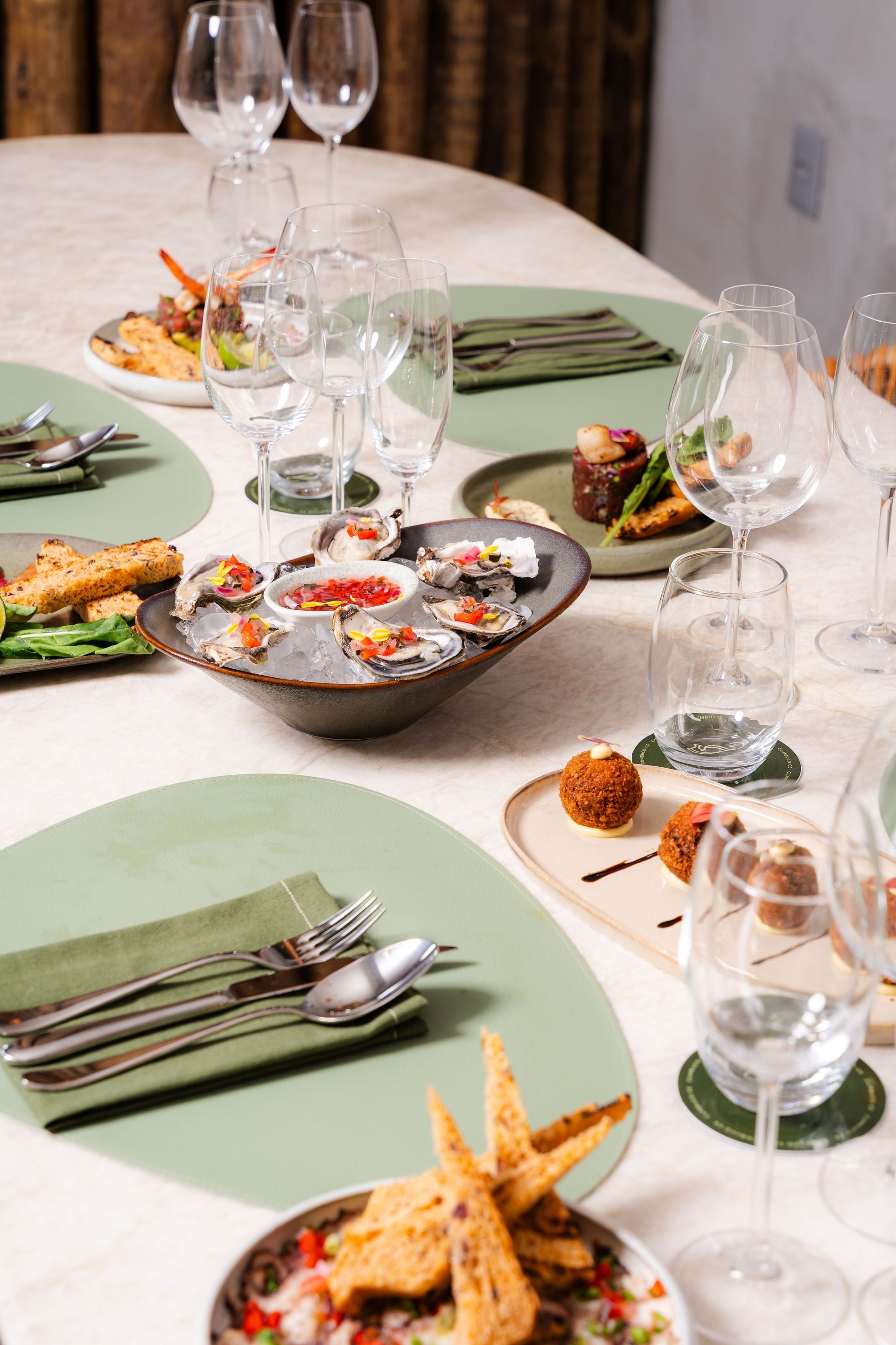 Elegante mesa de comedor con servilletas verdes, copas de vino y una variedad de aperitivos sobre mantel blanco.